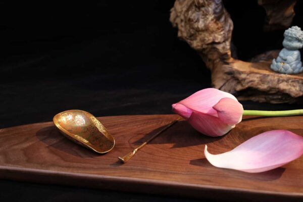 Artistic still life with a vintage golden tea scoop, brass spoon, and pink lotus flower on a walnut wood platter.
