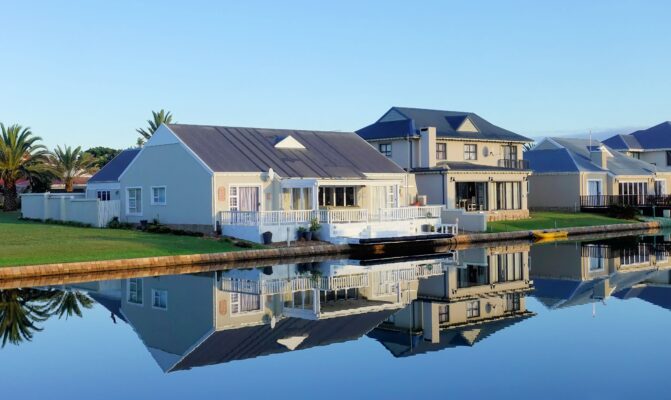 Luxury modern homes reflecting on a peaceful, clear lake during daylight, showcasing architectural beauty
