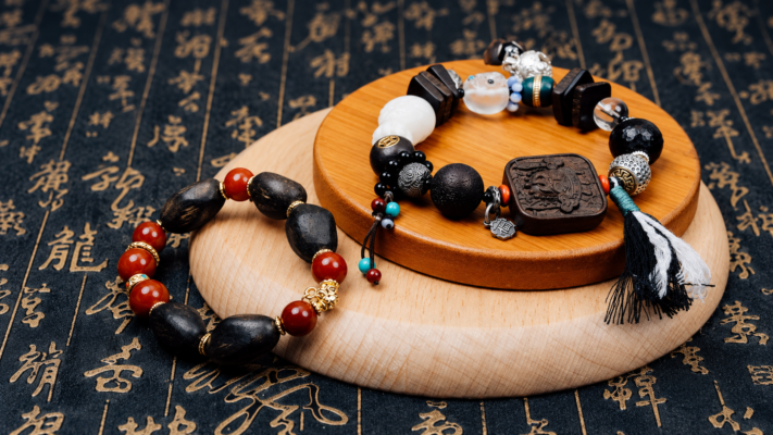 Two agarwood bracelet designs displayed on wooden stands over dark Chinese calligraphy fabric
