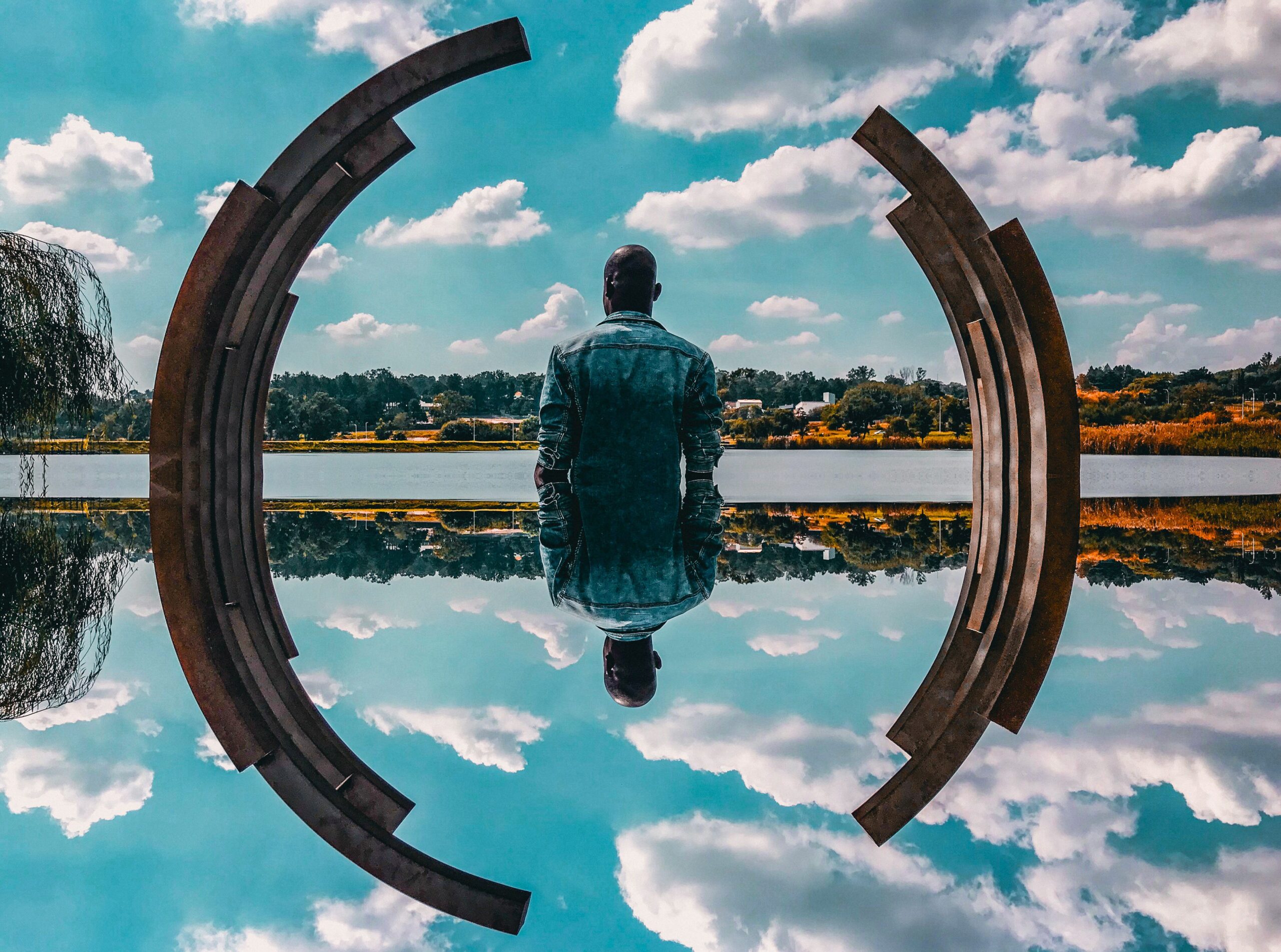 Seated person reflected in water beside a curved sculpture